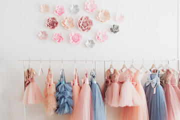 Beautiful dressy lush pink and blue dresses for girls on hangers at the background of white wall. Kids dresses with feathers for prom and holiday.