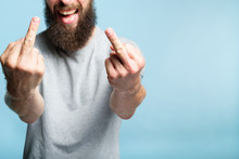 Bearded Man Showing Middle Fingers. Provocation Defiance Attitude And Offensive Indecent Behavior Concept. Cropped Shot Of A Male Torso On Blue Background. Casual Hipster In Grey T-shirt.