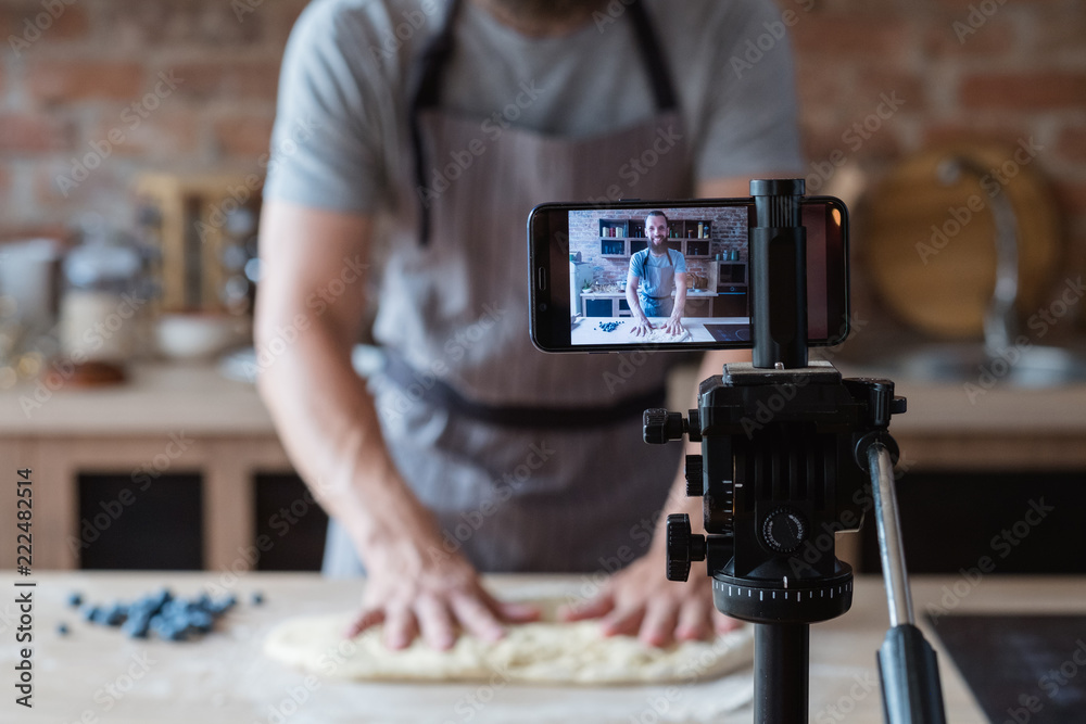 baker online courses. food preparing and culinary training class concept. smiling bearded chef kneading dough in the kitchen and shooting video of himself using mobile phone on a tripod. - obrazy, fototapety, plakaty 