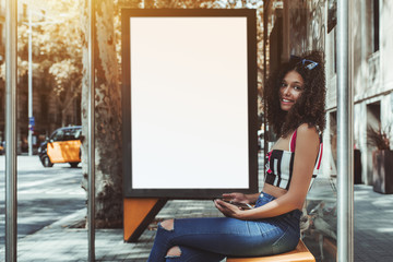 Wall Mural - A cheerful curly cute girl is sitting on the metal bench of an urban bus stop, holding smartphone while waiting for her transport, with blank white banner placeholder mock-up behind for your advert