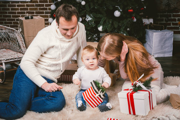 Wall Mural - Family celebratory Christmas evening. Family three Caucasian people sitting under coniferous tree Christmas tree on floor of carpet wool. A child holds box with gift, parents young stylishly dressed