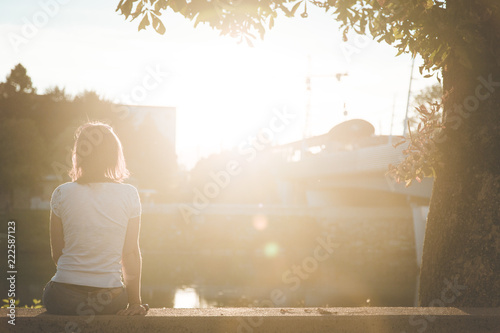 Fotografía Junge Frau sitzt in urbaner Gegend auf Mauer und blickt dem Sonnenuntergang entg