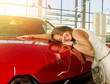 Dream about car. Gorgeous smiling woman hugging lies on the hood of new red car in the dealership.