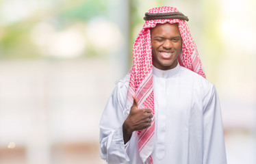 Wall Mural - Young arabic african man wearing traditional keffiyeh over isolated background doing happy thumbs up gesture with hand. Approving expression looking at the camera with showing success.