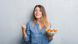 Young adult woman over grey grunge wall eating healthy tomato salad screaming proud and celebrating victory and success very excited, cheering emotion