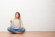 © Krakenimages.com - Young adult woman sitting on the floor over white brick wall at home showing and pointing up with finger number one while smiling confident and happy.