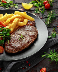 Grilled sirloin steak with potato fries and vegetables, tomato salad in a black plate. rustic table