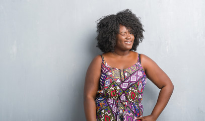 Wall Mural - Young african american woman over grey grunge wall wearing colorful dress winking looking at the camera with sexy expression, cheerful and happy face.