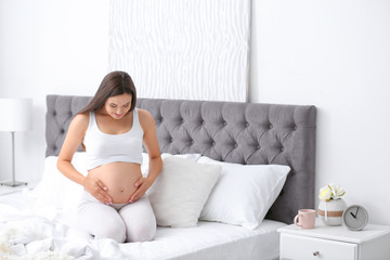 Wall Mural - Young beautiful pregnant woman sitting on bed and touching her belly at home