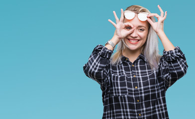 Young blonde woman wearing sunglasses over isolated background doing ok gesture with hand smiling, eye looking through fingers with happy face.