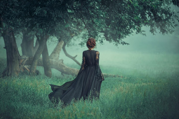 girl with a red hair walking along from dark forest, wearing long black dress with trailer which is 