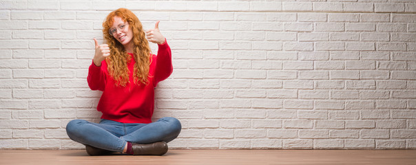 Poster - Young redhead woman sitting over brick wall approving doing positive gesture with hand, thumbs up smiling and happy for success. Looking at the camera, winner gesture.