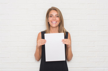 Beautiful young woman over white brick wall holding blank paper sheet with a happy face standing and smiling with a confident smile showing teeth