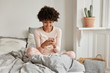 © WHstudio Leushin N - Excited delighted dark skinned housewife enjoys rest in bed, watches video via media app, has spare time, poses at comfortable bed on bedclothes, dressed in pyjamas, enjoys online marketing.