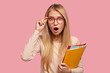 Emotional bugged eyed young blonde girl cant believe in sudden news, opens mouth widely, expresses wonder, wears transparent glasses, holds textbooks, stands over pink background. Omg concept