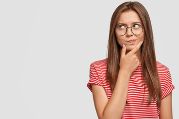 Wall Mural - Photo of unsure doubtful young woman holds chin, looks right doubtfully, reconsiders something, feels hesitant, dressed in casual striped t shirt, poses against white background with copy space