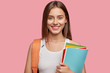 Horizontal shot of pleasant looking cheerful European girl with dark straight hair, dressed in casual white t shirt, carries rucksack and textbooks, isolated over pink background. Emotions, learning