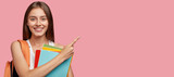 Fototapeta  - Studio shot of positive Caucasian woman with shining smile, dark straight hair, poins at upper right corner, shows free space for your advertisement, ready for school, isolated over pink background