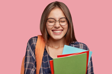 Wall Mural - Horizontal shot of cheerful Caucasian woman with broad smile, keeps eyes closed, being in high spirit after lectures or classes, has orange rucksack, stands indoor. Positive emotions concept