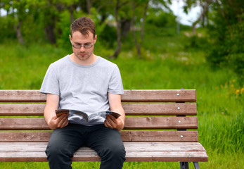 The young caucasian man is wearing casual clothes reading a magazine in a park. European male model, 27 years old, with glasses is sitting on a wood bench in outdoors.