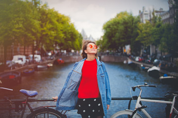 Wall Mural - Young girl in red sweater and orange sunglasses with backpack at bridge in Amsterdam street. Holland, Netherlands. Autumn season