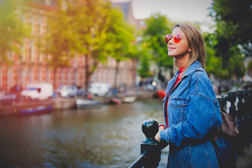 Wall Mural - Young girl in red sweater and orange sunglasses with backpack at bridge in Amsterdam street. Holland, Netherlands. Autumn season