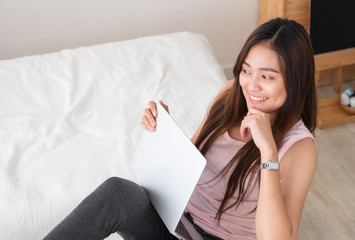 Poster - Asian female freelancer smile and holding laptop with rest leg on table in bedroom at home.Work at home concept.work from home concept.