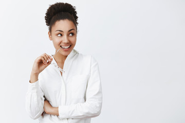 Girl intending to go and dinner with charming man from next office. Intrigued good-looking african woman in white shirt, gazing right and smiling broadly, biting rim of glasses from curiosity