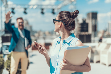 Poster - Hey, I am here. Positive bearded male gesticulating hands while telling with girl. She turning back to camera and looking at him. Female holding mobile and documents with information outdoor