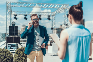 Poster - Laughing bearded male telling on contemporary cellphone while looking at camera. He keeping laptop while standing opposite lady with documents