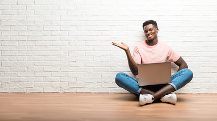 Wall Mural - Afro american man sitting on the floor with his laptop holding copyspace imaginary on the palm