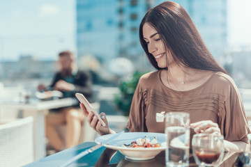 Poster - Portrait of beaming female typing in phone while sitting at table outside. She eating delicious cuisine
