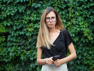 Portrait of young beautiful long hair woman wearing black blouse, holding mobile phone at summer green park against ivy background.