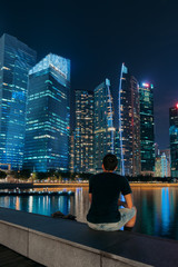 Singapore city skyline. Man is sitting near business district view. Downtown reflected in water at night in Marina Bay. Travel cityscape