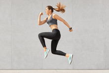 Active Woman Being Full Of Energy, Jumps High In Air, Wears Sportsclothes, Prepares For Sport Competitions, Isolated Over Grey Concrete Wall. Female Trainer Busy With Training. Gymnastics Concept