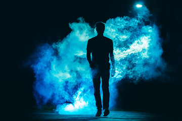 The male walking in the blue smoke on the dark background. evening night time