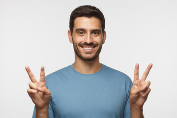 Indoor portrait of young male isolated on gray background with optimistic smile, showing victory gesture with both hands, looking friendly. V sign