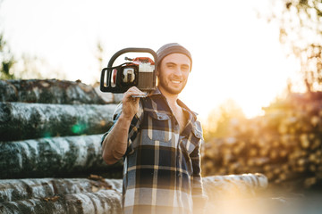 Bearded strong lumberjack wearing plaid shirt hold in hand chainsaw for work on sawmill