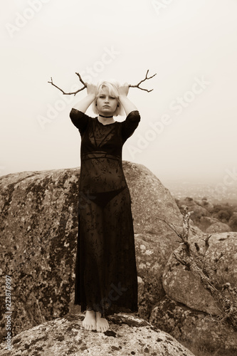 Fotografía Artistic photo of a blond girl in black dress and sinister aesthetics on the rocks in the countryside on a cloudy day