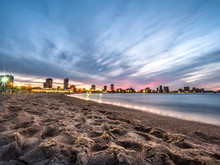 Beautiful Sunset Photograph Of The Beach And Sand  Along The Shoreline Of Lake Michigan At North Avenue Beach In Chicago With Colorful Pink And Blue Sky And Lights Near The Beach House Beyond.