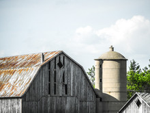 Beautiful Photograph Of A Weathered Old Barn With Rusty Tin Roof And Concrete Silo With White Fluffy Clouds In The Sky Above.