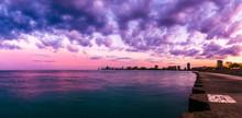 Beautiful Panorama Photograph Of The Chicago Skyline At Sunset With Fluffy Purple And Pink Clouds In The Sky Above And Blue Water Below Along The Concrete Shoreline At Montrose Beach.