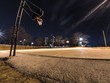 Wide angle photograph of a concrete exterior basketball court with hoops at either end, street lights with residential highrise apartments in the distance in the dark night sky in Chicago.