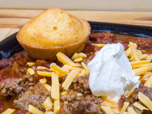 Homemade Golden Brown Cornbread Muffin Placed On Top Of A Hot Bowl Of Cheese And Sour Cream Topped Chili On A Butcher Block Counter.