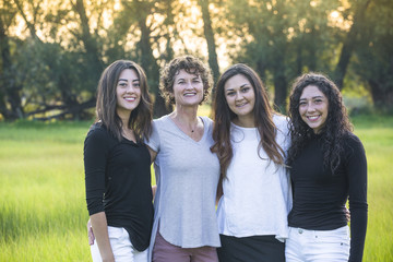 Wall Mural - Outdoor Portrait of a beautiful family, a mother and her daughters. Horizontal portrait of beautiful hispanic women standing together in a grass field with the sun at their backs