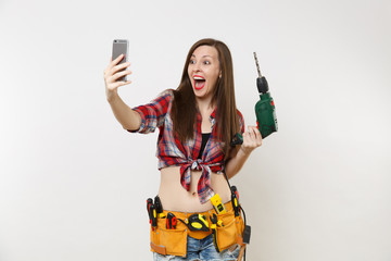 Wall Mural - Strong excited handyman woman in shirt, kit tools belt full of instruments doing selfie on mobile phone with power electric drill isolated on white background. Female in male work. Renovation concept.