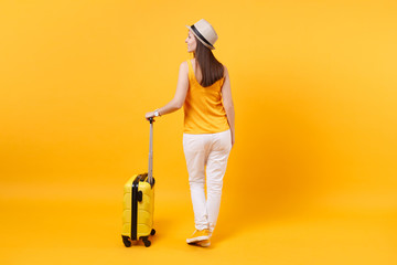 Back view tourist woman in summer casual clothes, hat with suitcase isolated on yellow orange background. Female passenger traveling abroad to travel on weekends getaway. Air flight journey concept.