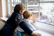 Happy adorable kid boy and cute baby girl sitting near window and looking outside on snow on Christmas day or morning