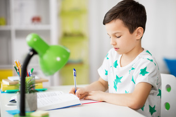 Wall Mural - education, childhood and school concept - boy with book writing to notebook at home