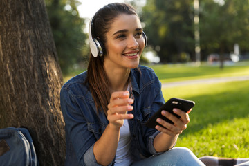 Poster - Beautiful young woman student in the park listening music with headphones using mobile phone.